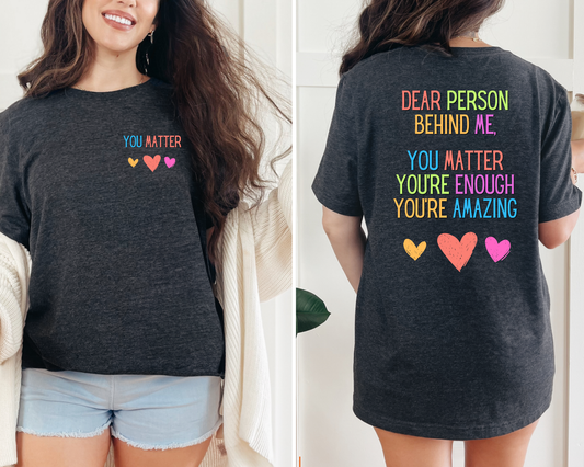 A graphic T-shirt with the message 'YOU MATTER' printed in colorful letters on the front and a message 'DEAR PERSON BEHIND ME, YOU MATTER ENOUGH, YOU'RE AMAZING' on the back, displayed on a person.